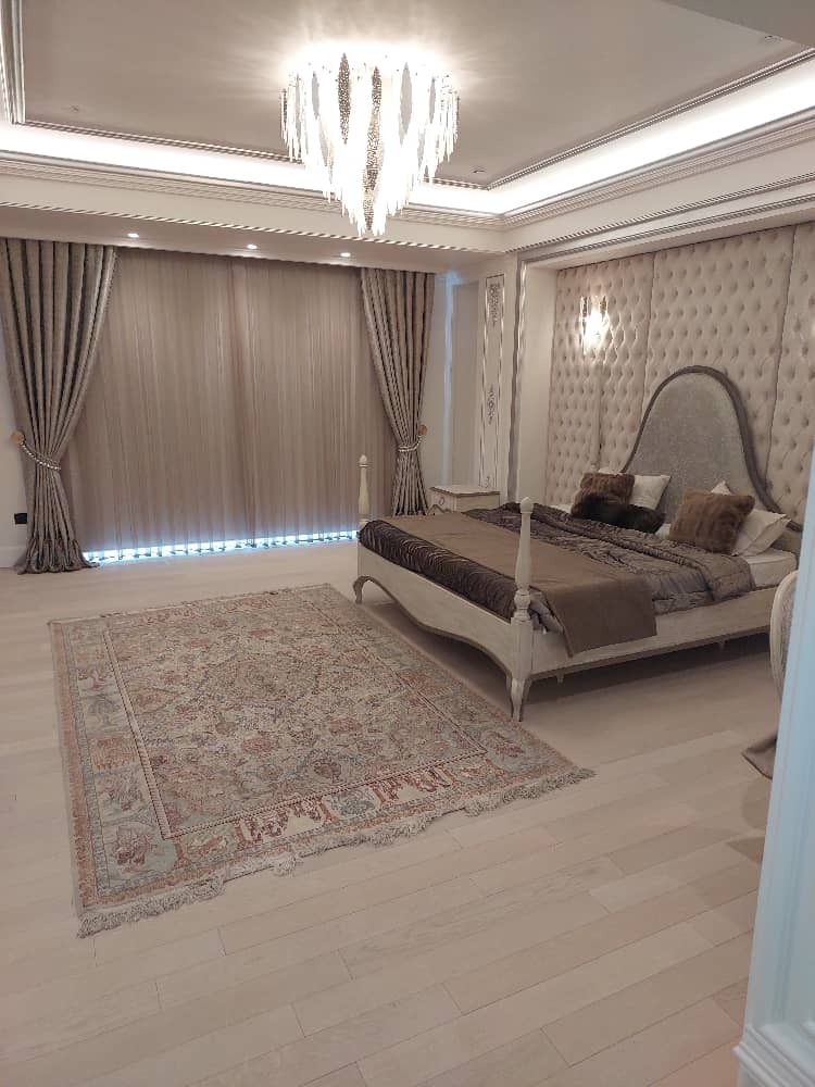 Modern luxury bedroom featuring padded wall design, chandelier lighting, full-length curtains, and Persian rug